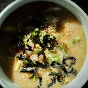 A bowl of creamy vegan rice cake soup topped with seaweed strips, chopped green onions, and sesame seeds, with dumplings. The dish is set against a dark background.